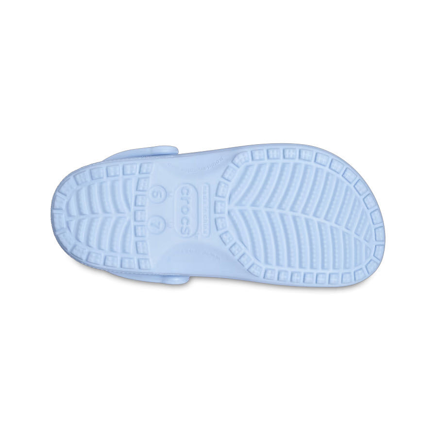 The image displays the sole of a CROCS CLASSIC CLOG BLUE CALCITE - ADULTS, featuring tread patterns and the Crocs logo, emphasizing the comfort shoe&#39;s unique design.