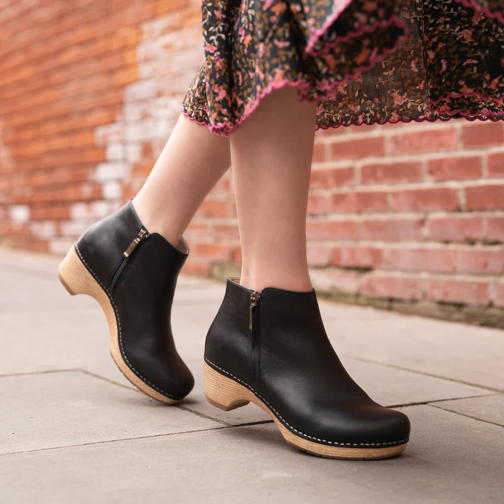 A blonde wearing a floral skirt and Dansko Lizanne Black short booties with wooden soles walks on a paved surface beside a red brick wall.