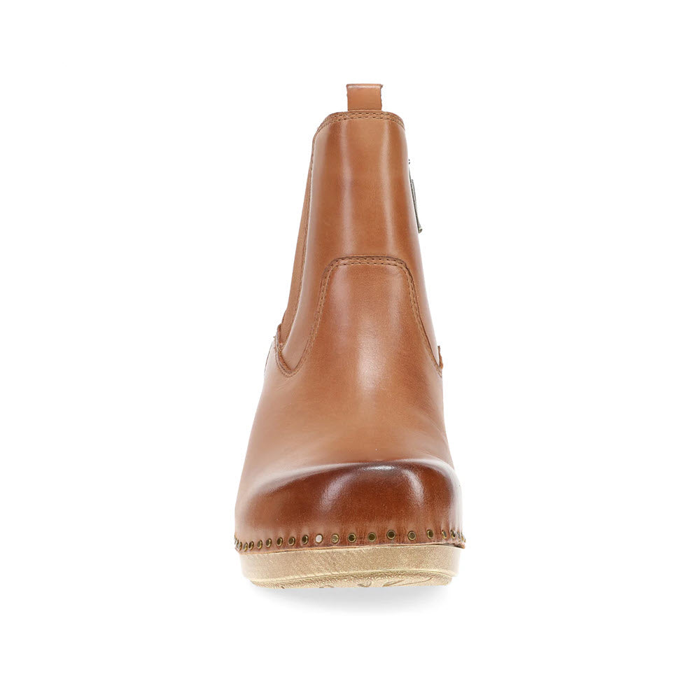 Front view of the Dansko Shayna Luggage heeled boot in tan leather, showcasing a rounded toe, elastic side panels, and a light-colored wooden sole accented with small decorative studs along the edges. This statement piece from Dansko seamlessly blends style and comfort.