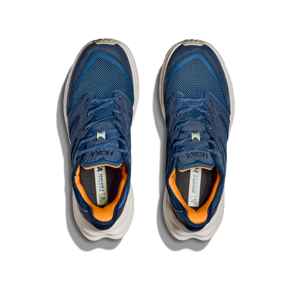 A pair of HOKA ANACAPA 2 FREEDOM FOGGY NIGHT/OAT MILK running shoes, with blue uppers, white soles, and an orange inner lining, viewed from above to highlight the HOKA geometry for optimal performance.