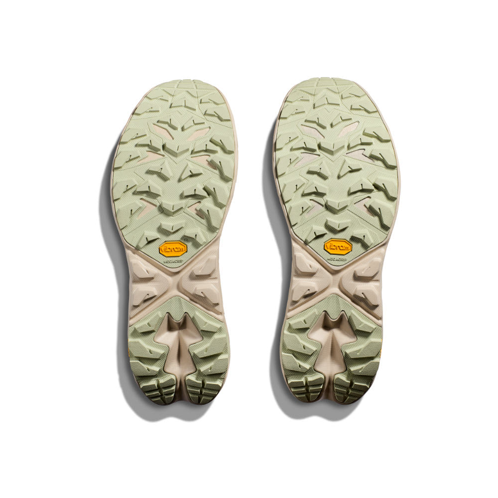 Two light green Hoka ANACAPA 2 FREEDOM FOGGY NIGHT/OAT MILK men&#39;s shoe soles with visible treads and a yellow logo on each, displayed on a white background, showcasing the durability of Vibram Megagrip technology.