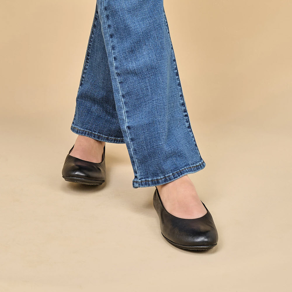 A person wearing blue jeans and the chic Dansko Mollie black ballet flats stands on a beige background.