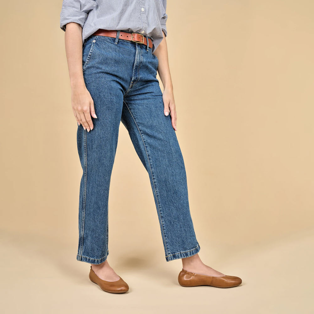 A person wearing a light blue shirt, blue jeans, a brown belt, and chic Dansko Mollie Luggage - Womens flats stands against a beige background.