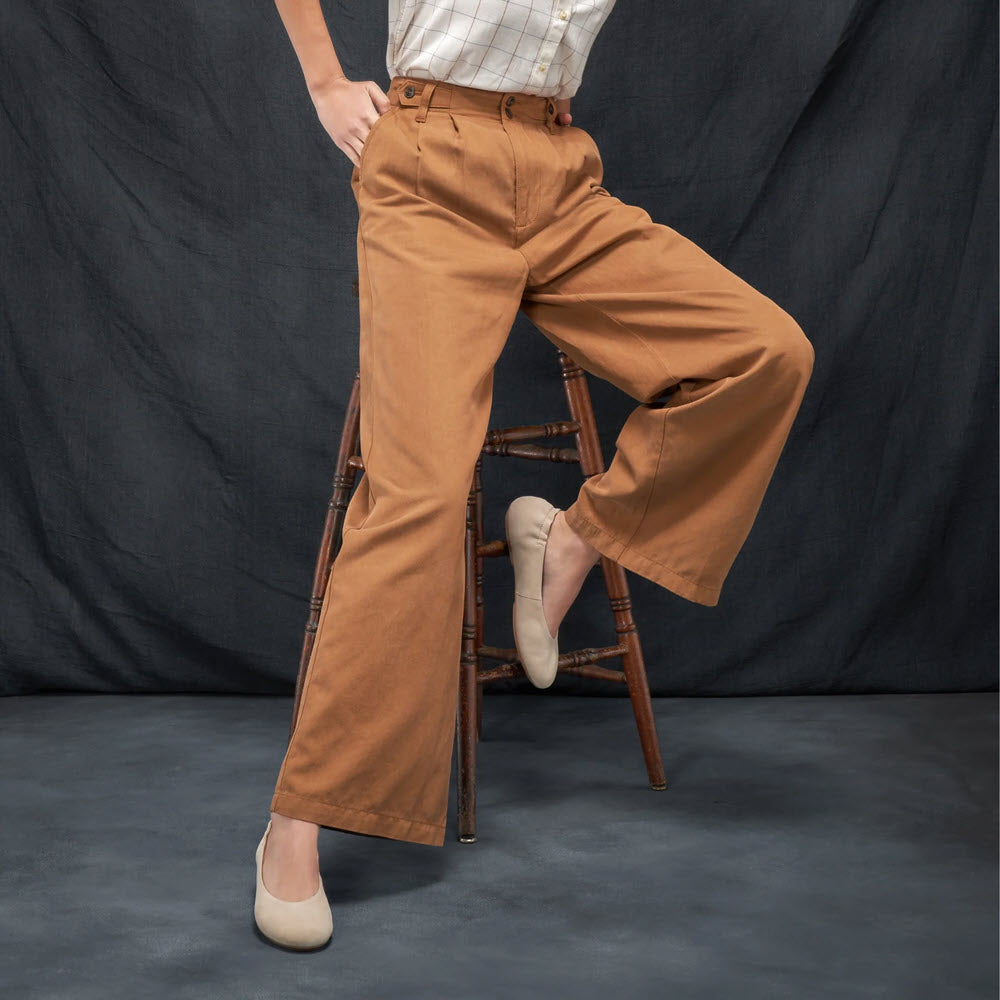 Person in a checked shirt and brown wide-leg pants sits on a wooden stool against a dark fabric backdrop. Only the lower body is visible, showcasing chic comfort with beige Dansko DANSKO MOLLIE SAND - WOMENS flats.