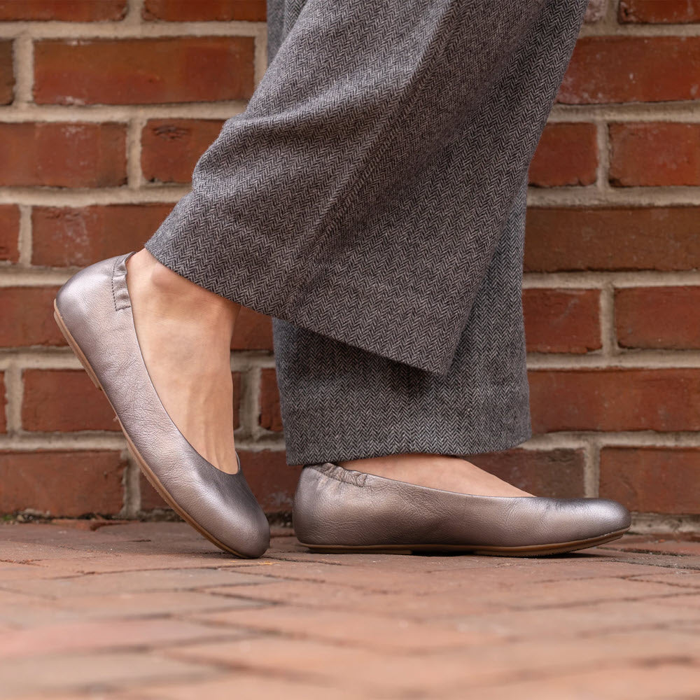 Person wearing grey Dansko DANSKO MOLLIE PEWTER - WOMENS and grey herringbone pants stands on a brick-paved surface in front of a brick wall.