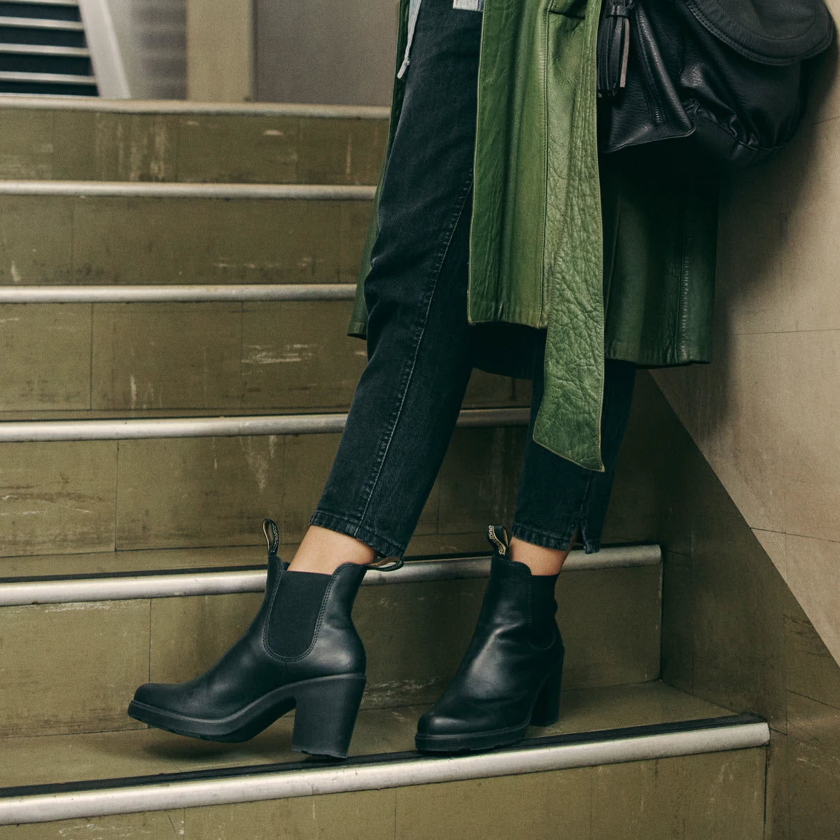 Person wearing a green coat, black jeans, and Blundstone BLUNDSTONE 2365 BLOCK HEEL BLACK - WOMENS stands on a set of stairs.