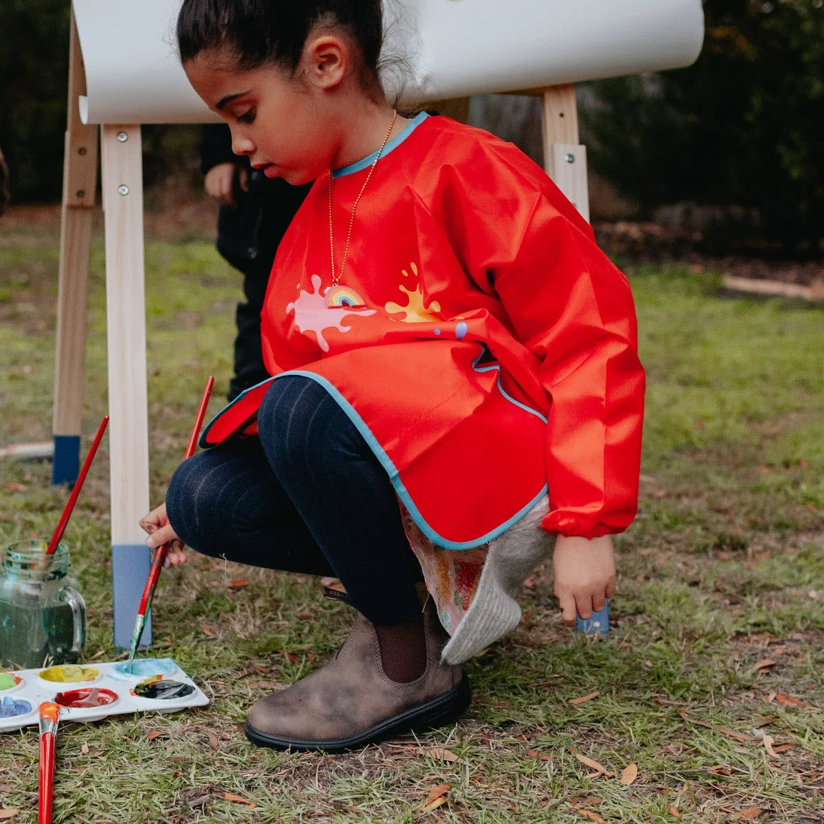 A child kneels on the grass, clad in a red smock with paint splatter designs and Blundstone 565 Chelsea Boots in Rustic Brown for added durability. Holding a paintbrush, they are surrounded by painting materials on the ground, with an easel in the background.