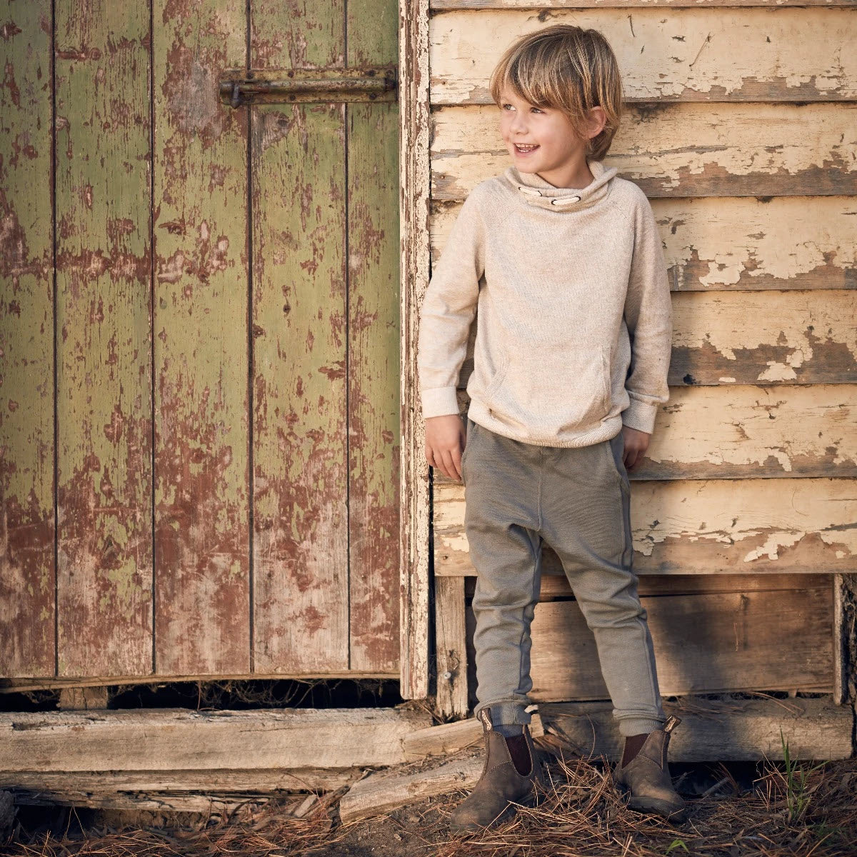 A child stands smiling beside a weathered wooden wall and door. The child wears a light sweater, green pants, and durable Blundstone BLUNDSTONE 565 CHELSEA BOOTS RUSTIC BROWN - KIDS known for their excellent shock absorption.