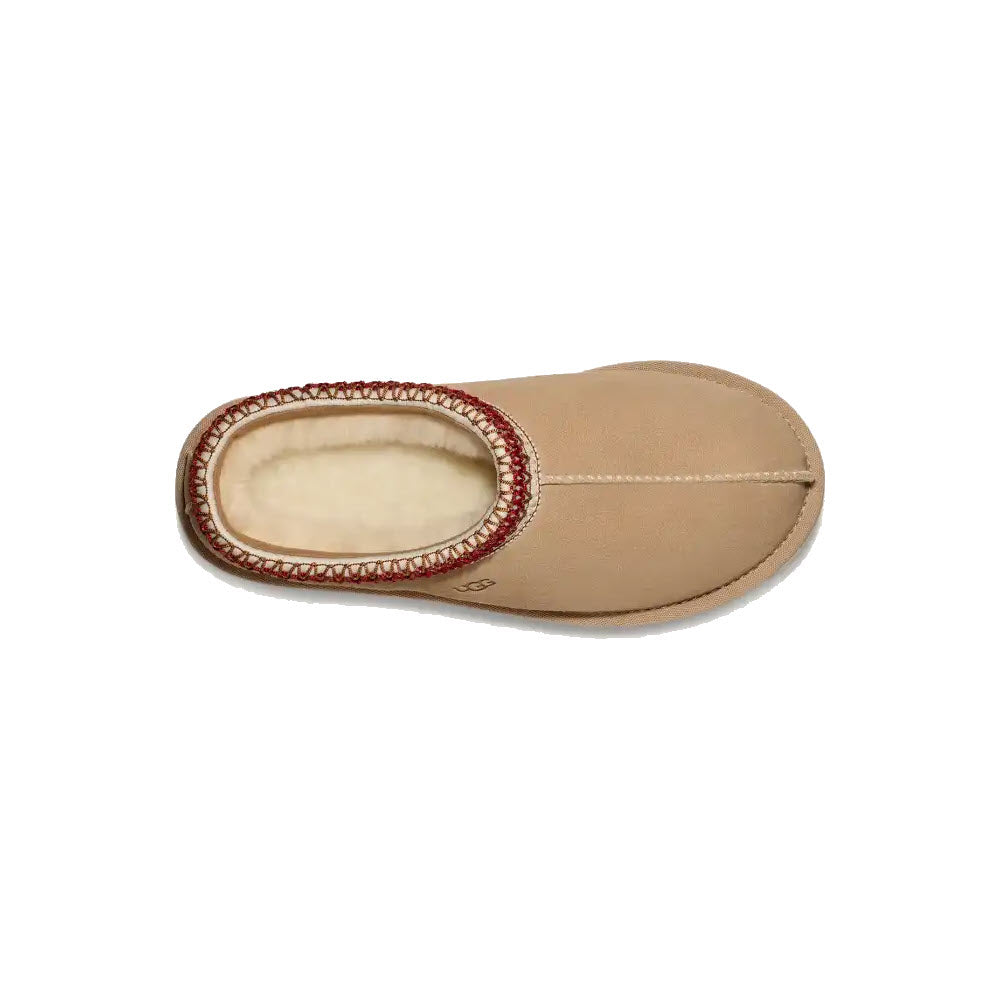 The UGG TASMAN SAND/DARK CHERRY - WOMENS house slipper by Ugg features a tan color with a soft interior lining and red stitching around the collar, viewed from above. Crafted for durability and comfort, this timeless piece ensures that your feet stay cozy all day long.