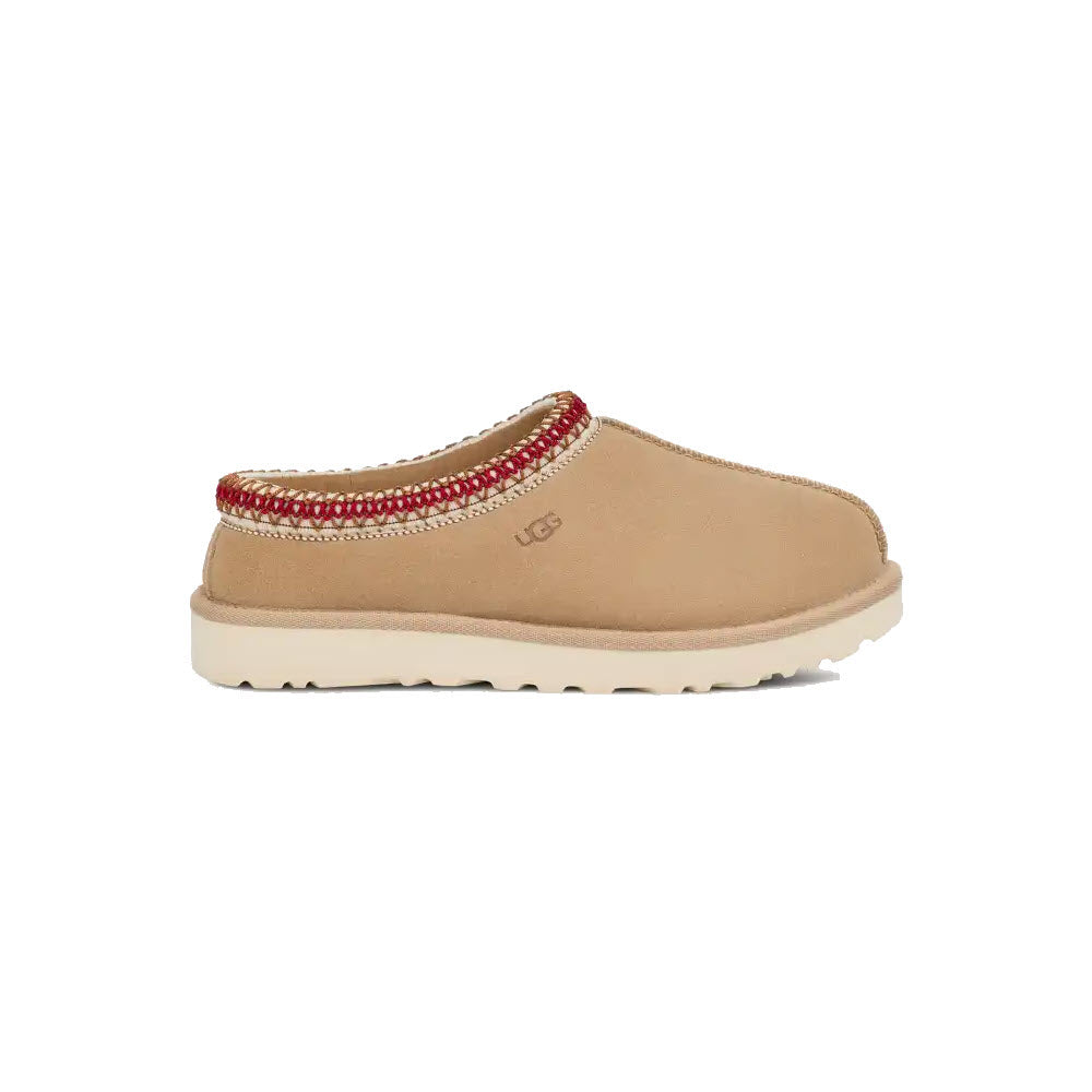 Side view of the UGG Tasman Sand/Dark Cherry - Women&#39;s slip-on shoe, featuring a tan exterior with a red-trimmed opening and a beige rubber sole. The durable shoe showcases the &quot;UGG&quot; logo on the side, merging timeless style with practical design.