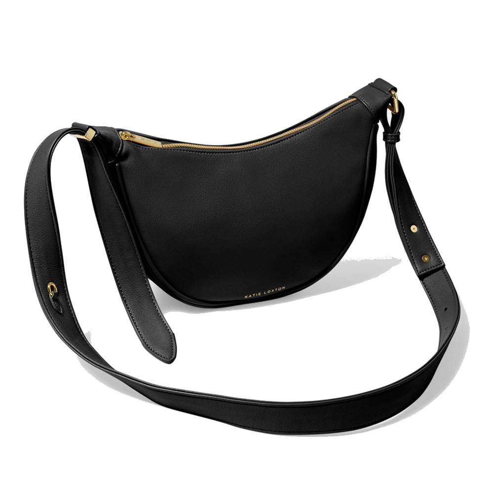 The Katie Loxton Harley Sling Saddle Bag Black, crafted from black vegan leather, features gold-tone hardware and an adjustable crossbody strap, shown on a white background.