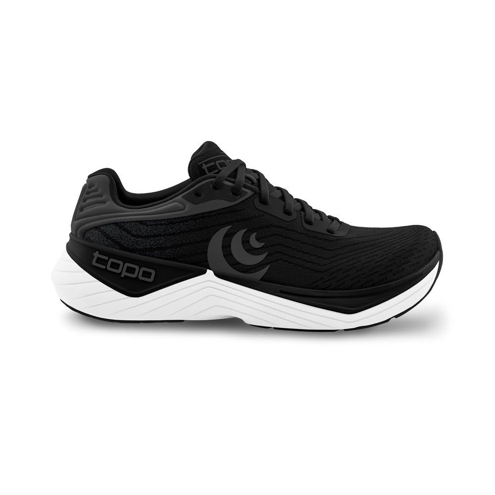 The TOPO ULTRAFLY 5 BLACK/WHITE - WOMENS sneaker is a black athletic shoe featuring a white sole and the brand name "Topo" on both the side and tongue. It boasts a minimalist design with textured details, a cushioned heel, and light pronation support to ensure a stable run.