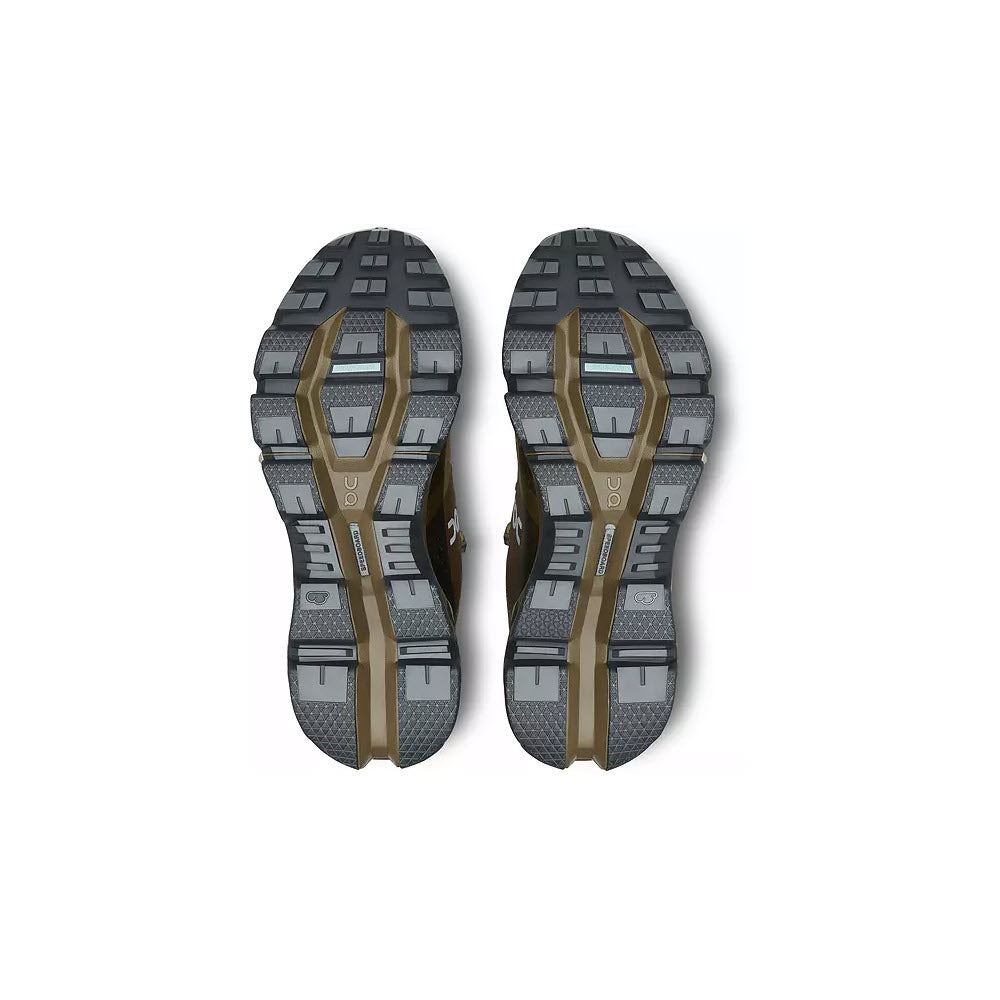 The image displays the outsoles of the ON CLOUDWANDER WATERPROOF HUNTER/SAFARI shoes by On Running. These lightweight women&#39;s athletic shoes feature a combination of black and grey rubber treads with olive green accents. The soles, designed for outdoor performance, have a segmented pattern that provides superior grip.