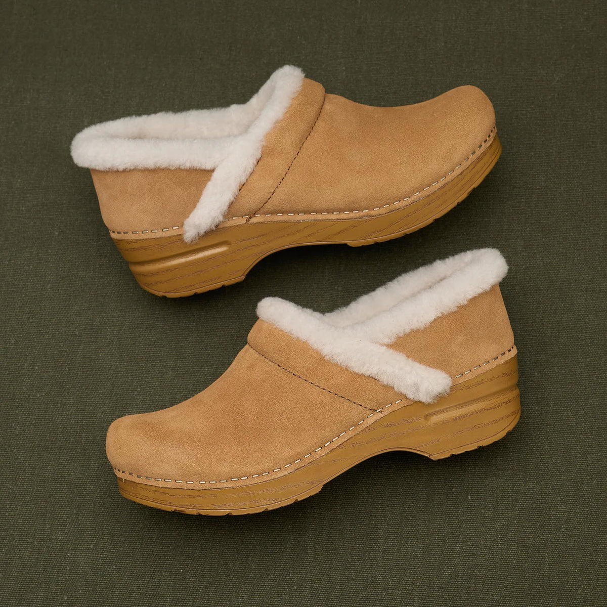 The Dansko Professional Cozy Suede clogs for women, crafted in tan suede with a contoured footbed and white fleece lining, featuring wooden soles, gracefully rest on a dark green surface.