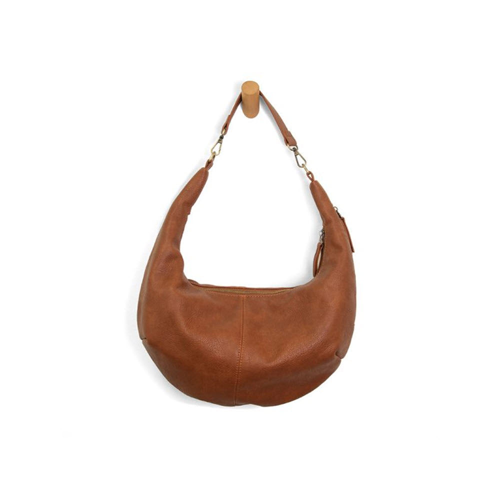 The JOY SUSAN RHEA SLOUCHY SHOULDER BAG SADDLE by Joy Susan hangs by its strap on a wooden peg against a white background. The tumbled vegan leather gives it a chic, timeless appeal perfect for everyday wear.