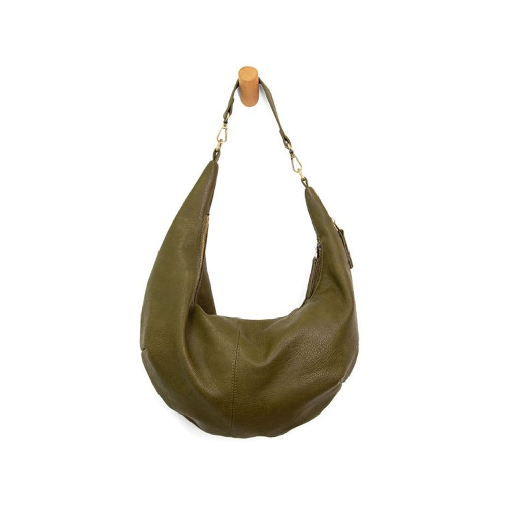 The JOY SUSAN RHEA SLOUCHY SHOULDER BAG in olive, by Joy Susan, with its crescent shape and gold hardware, is crafted from tumbled vegan leather and hangs stylishly on a wooden peg against a pristine white background.